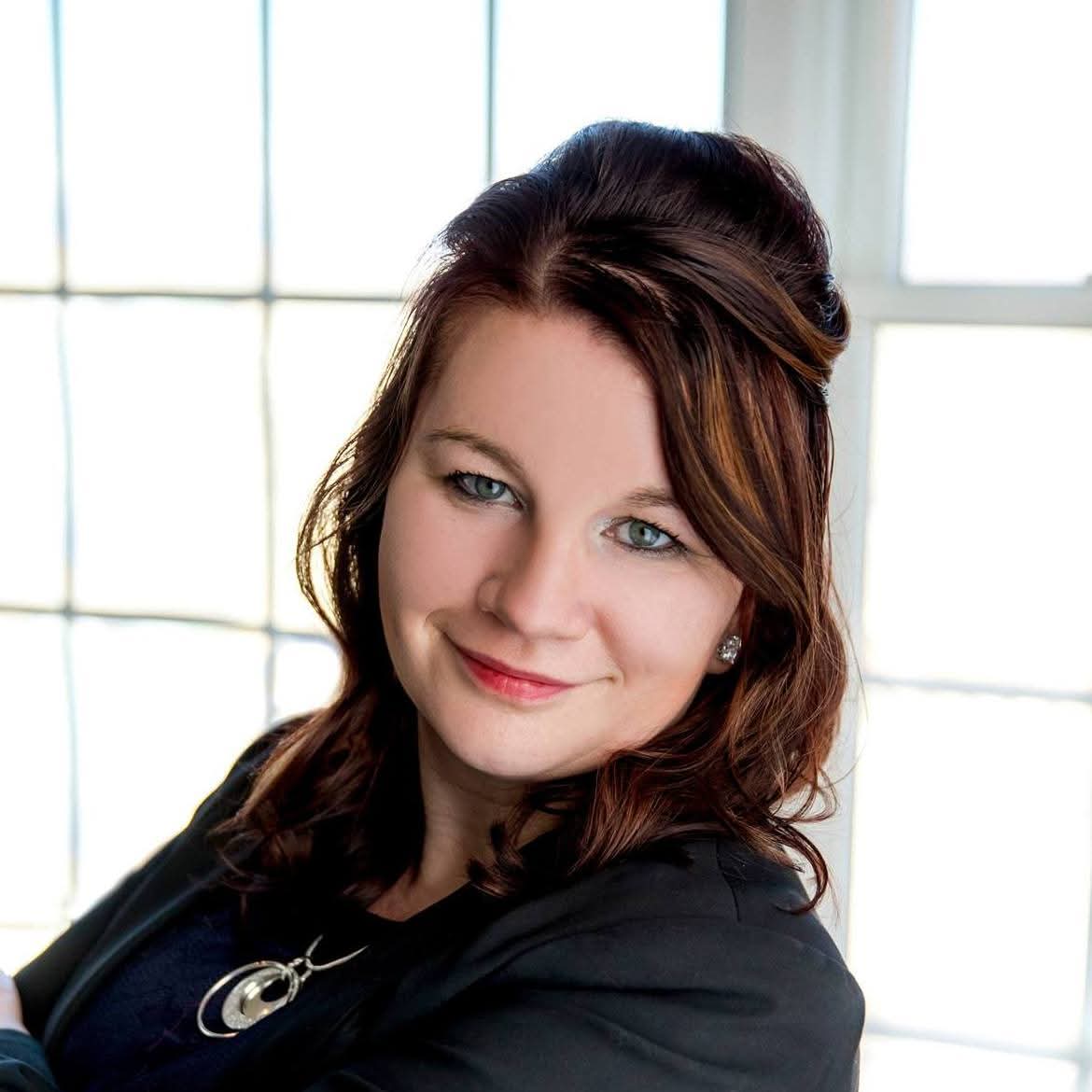 A woman with shoulder-length brown hair smiles confidently, wearing a black blazer and a silver pendant necklace, with a bright window background.