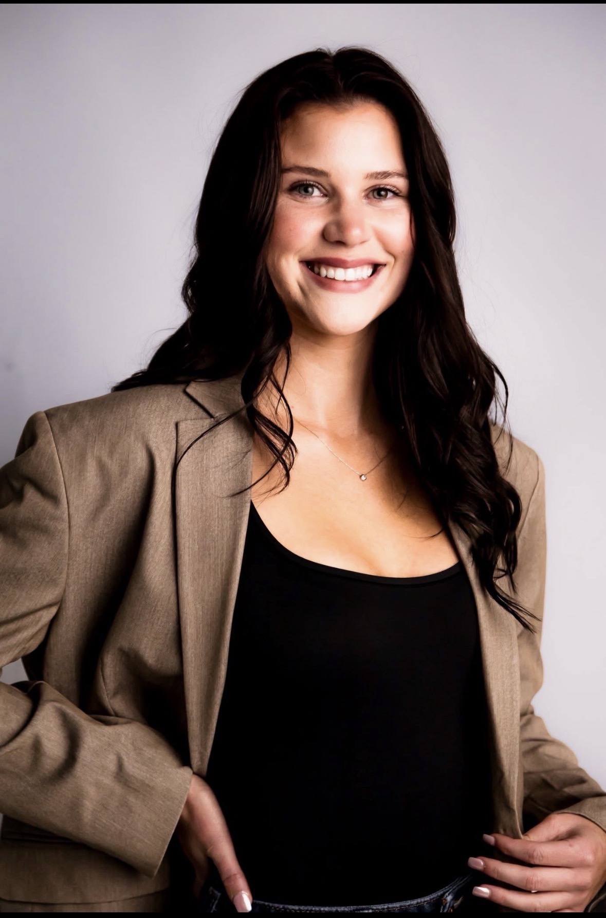 A smiling woman with long dark hair wearing a tan blazer over a black top, posing confidently with one hand on her hip.