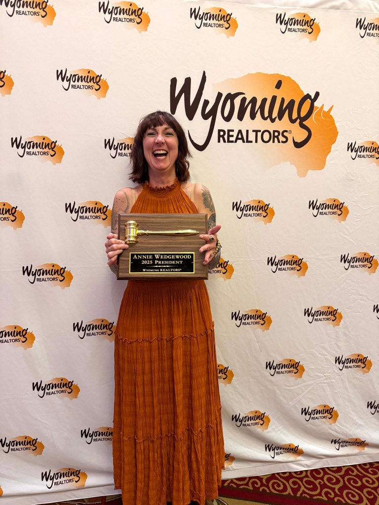 A person wearing an orange dress smiles broadly while holding an award in front of a 'Wyoming Realtors' backdrop.